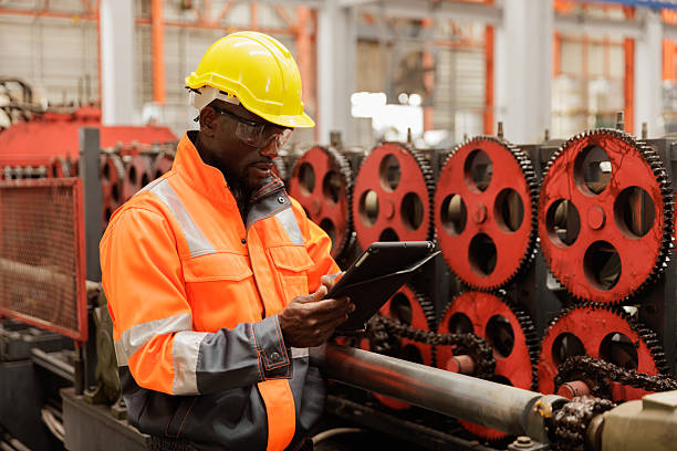 Industrial engineer wearing safety helmet and high-visibility uniform checks production data on a tablet while standing beside mechanical gears inside a manufacturing plant. Concept of modern technology, factory inspection, and digital monitoring in industrial operations.